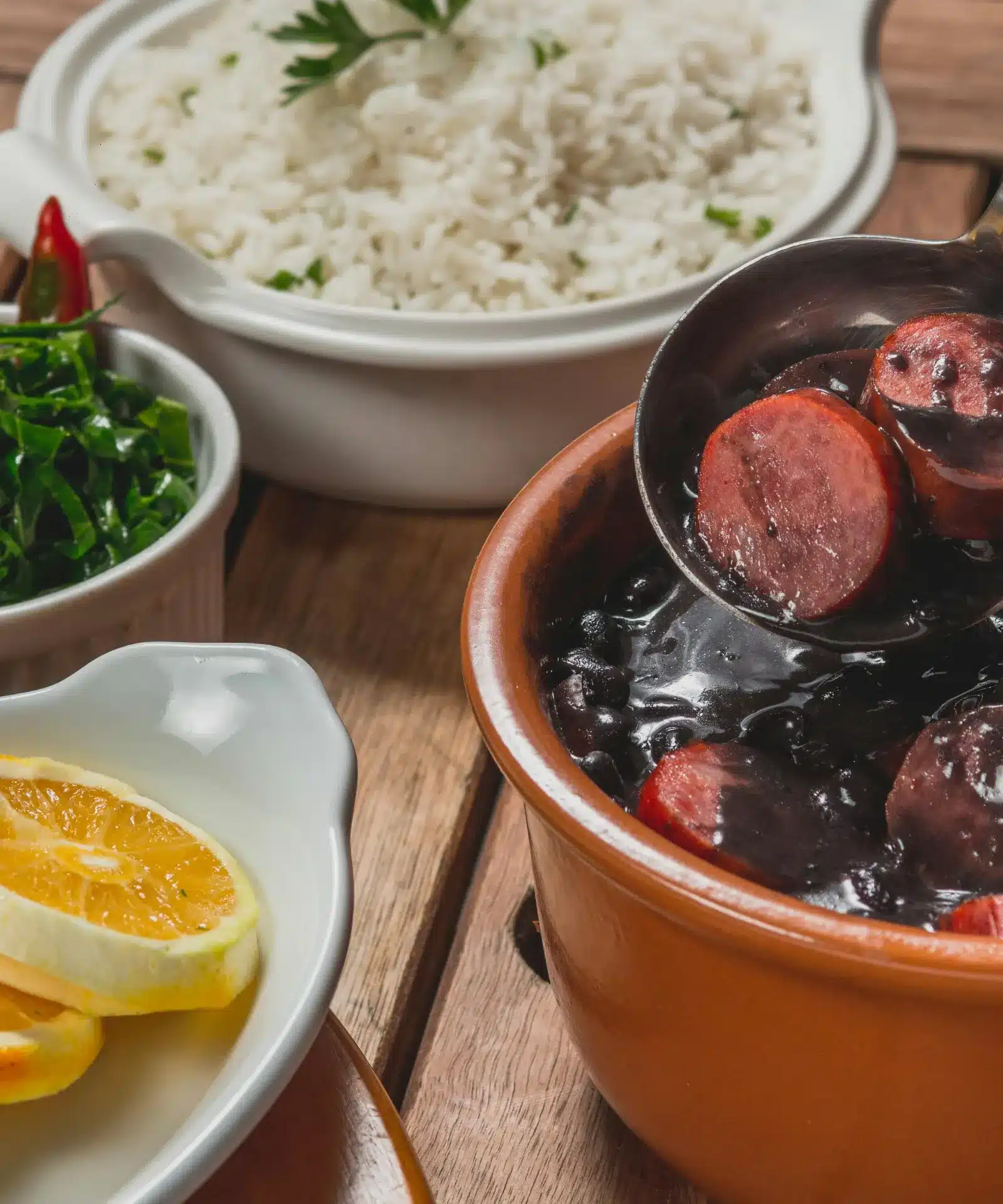 A served feijoada with black beans and sausage, alongside white rice, collard greens and orange slices.